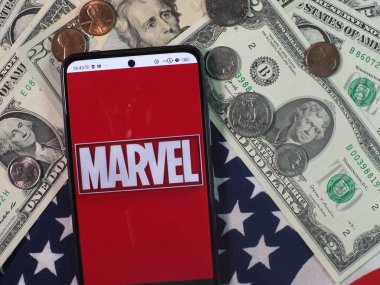 Bu fotoğraf illüstrasyonunda, Marvel Entertainment, LLC logosu arka planda ABD Doları banknotları ve bozuk paralarla bir akıllı telefonda görüntülenir.