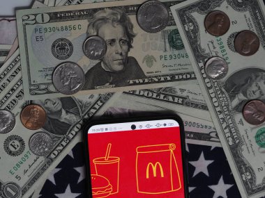 Bavyera, Almanya - 25 Aralık. 2023: Bu fotoğraf illüstrasyonunda, arka planda ABD Doları banknotları ve paraları olan bir akıllı telefonda McDonald 's Corporation logosu görülüyor. 