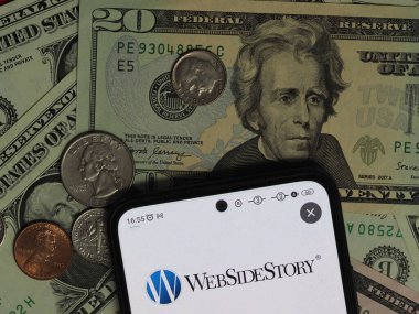 Bavyera, Almanya - 27 Aralık. 2023: Bu fotoğraf illüstrasyonunda, arka planda ABD Doları banknotları ve sikkeleri bulunan bir akıllı telefonda bir WebSideStory, Inc. logosu görülüyor. WebSideStory, Adobe Şirketi 'nin astı.