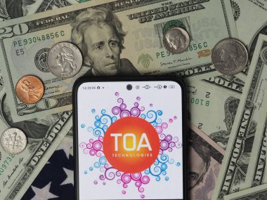 Bavyera, Almanya - 7 Ocak 2024: Bu fotoğraf illüstrasyonunda, arka planda ABD Doları banknotları ve madeni paraları olan bir TOA Teknolojileri logosu gösteriliyor.