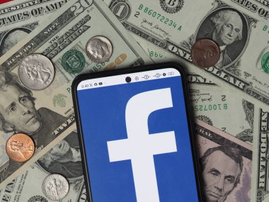 Bavyera, Almanya - 9 Ocak 2024: Bu fotoğraf illüstrasyonunda, arka planda ABD Doları banknotları ve paraları bulunan bir akıllı telefonda bir Facebook logosu görülüyor.