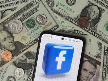 Bavyera, Almanya - 9 Ocak 2024: Bu fotoğraf illüstrasyonunda, arka planda ABD Doları banknotları ve paraları bulunan bir akıllı telefonda bir Facebook logosu görülüyor.