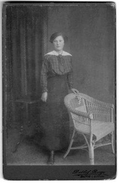 Beyaz dantelli yakalı ve kravatlı bir kadının stüdyo portresi, hasır bir sandalyenin ve küçük tahta bir masanın yanında duruyor, fotoğraf stüdyosu Detlef Ruge, Burg (Dithmarschen), Schleswig-Holstein, Almanya. Tarih bilinmiyor, yaklaşık olarak.