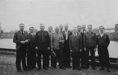 Antika bir fotoğraf, bir grup beyaz adamı gösteriyor, bazıları üniformalı ve diğerleri de takım elbiseli, bir limanın önünde poz veriyorlar. Hamburg, Almanya. 16 Eylül 1946.