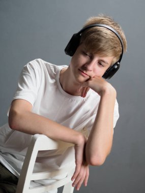 Beyaz tişörtlü, gri bir duvara yaslanmış beyaz bir sandalyede beyaz kulaklıklı, mavi gözlü, sarışın, düşünceli bir gencin stüdyo portresi.
