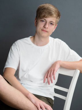 Beyaz tişörtlü, gri duvarda beyaz sandalyede oturan, beyaz gözlü, mavi gözlü sarışın bir gencin stüdyo portresi.