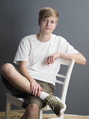 Beyaz tişörtlü, gri duvarda beyaz sandalyede oturan, beyaz gözlü, mavi gözlü sarışın bir gencin stüdyo portresi.