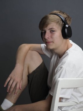 Beyaz tişörtlü, gri duvarlı beyaz sandalyede beyaz kulaklıklı, mavi gözlü sarışın bir gencin stüdyo portresi.