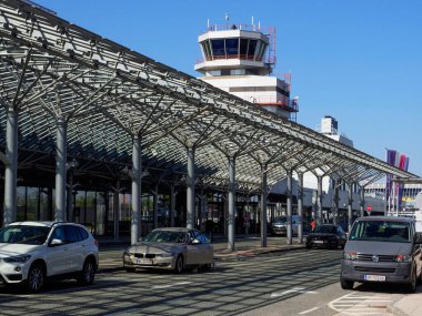 Horsching, Avusturya - 12 Ağustos 2025: Linz Havaalanı 'nın dış tarafı, sütunlar tarafından desteklenen eşsiz bir tepe örtüsü yapısı ile modern girişe sahiptir. Havaalanının kontrol kulesi açık mavi gökyüzüne karşı arka planda görünür. Arabalı bir yol.