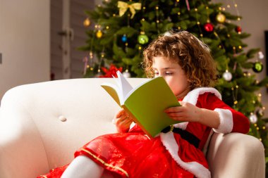 Güzel küçük kız güzel dekore edilmiş bir Noel ağacının yanındaki sandalyede oturmuş kitap okuyor.