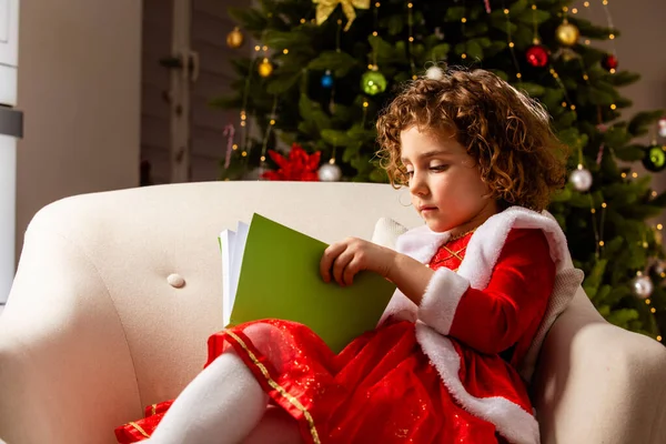 Krásná Holčička Sedí Židli Vedle Krásně Zdobeného Vánočního Stromečku Čte — Stock fotografie