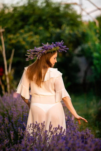 Woman in lavender wreath and whitre dress on the lavender field. Girl collects lavender.