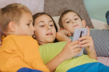 Çocuklar oyun odasında, battaniyenin altında yastıkların üzerinde akıllı telefonlarıyla oynuyorlar.