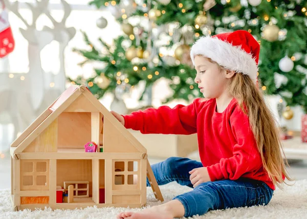 クリスマスの時間にサンタの帽子をかぶった小さな女の子が床に座って お祝いの木で飾られた新年の家で木製のおもちゃの家で遊んでいます かわいいです子供でクリスマス時間 — ストック写真