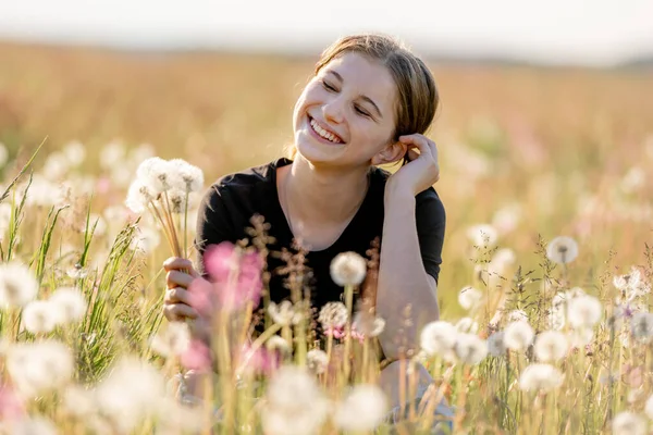 飛行タンポポと笑顔でフィールドに座っている可愛い女の子のティーンエイジャー 美しい若い女性モデルに自然で晴れた日の肖像画 — ストック写真