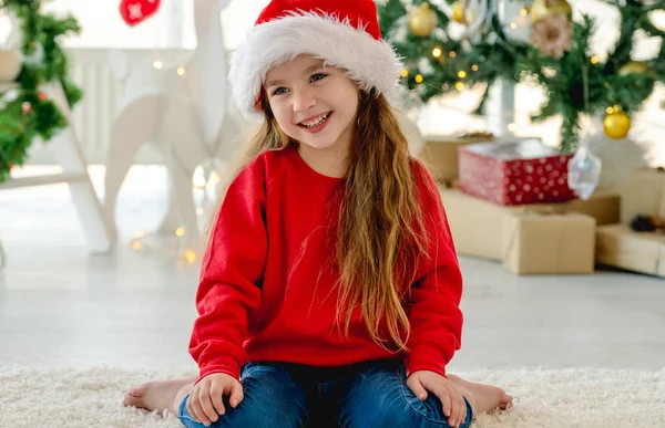 圣诞节快乐的小女孩坐在地板上 背对着装饰过的树在房间里笑 新年的时候 一个戴着圣诞礼帽的漂亮孩子在家里 — 图库照片