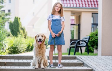 Yaz aylarında kotlu bir elbise giyen ve Golden Retriever köpeğiyle dışarıda kameraya bakan küçük bir kız. Şehirde safkan tüylü köpek evcil hayvanı olan güzel bir çocuk.