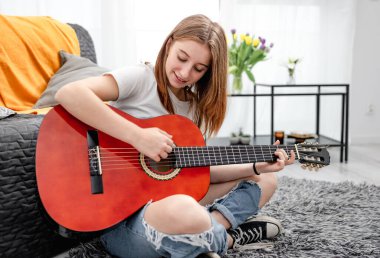 Evde gitar çalan genç bir kız yerde oturuyor. Müzisyen enstrümanıyla güzel bir gitarist.
