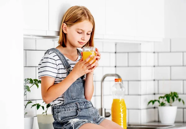 Preteen girl with orange juice bottle and glass sitting at kitchen table. Pretty child kid with vitamin fruit drink at home