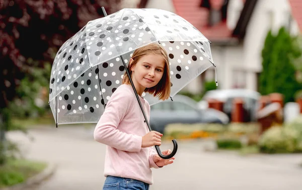 Preteen girl with umbrella looking at camera and smiling outdoors. Pretty child kid portrait in rainy day at street