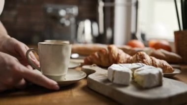 Girl takes coffee cappuccino cup for breakfast with croissants and brie cheese from table at kitchen. Tasty morning meal with espresso beverage and pastry