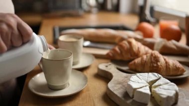 Girl put milk to coffee cappuccino cup for breakfast with croissants and brie cheese on table at kitchen. Tasty morning meal with creamy espresso beverage and pastry
