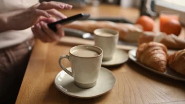 Girl chatting at smartphone and drink morning coffee cappuccino from cup. Woman with creamy espresso and mobile phone at kitchen