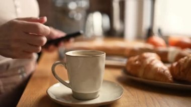 Girl chatting at smartphone and drink morning coffee cappuccino from cup. Woman with creamy espresso and mobile phone at kitchen