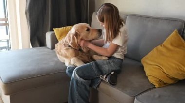 Preteen girl hugging golden retriever dog and smiling sitting on sofa at home. Beautiful child kid with labrador doggy pet indoors