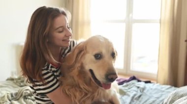 Güzel kız Golden Retriever köpeğine sarılıyor ve yatakta oturmuş gülümsüyor. Saf köpek labrador 'u olan genç bir kız evde kameraya bakıyor.
