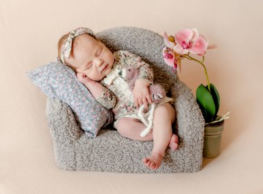 Cute newborn baby girl sleeping on tiny sofa with orchid flower decor studio portrait. Adorable little infant child kid napping