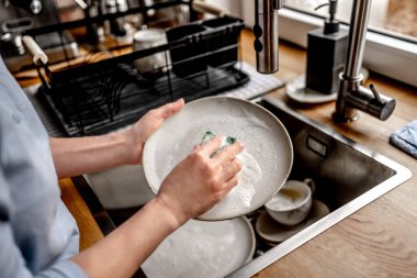 Girl washing plate dishes at kitchen at home with soap, foam and water. Woman cleaning utensil in sink