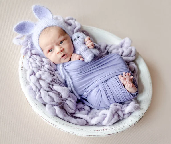 新生児の女の子ウサギの耳にウサギのおもちゃを保持し カメラを見てニット帽を着て 幼児子供嘘Swaddledでパープル生地スタジオポートレート — ストック写真