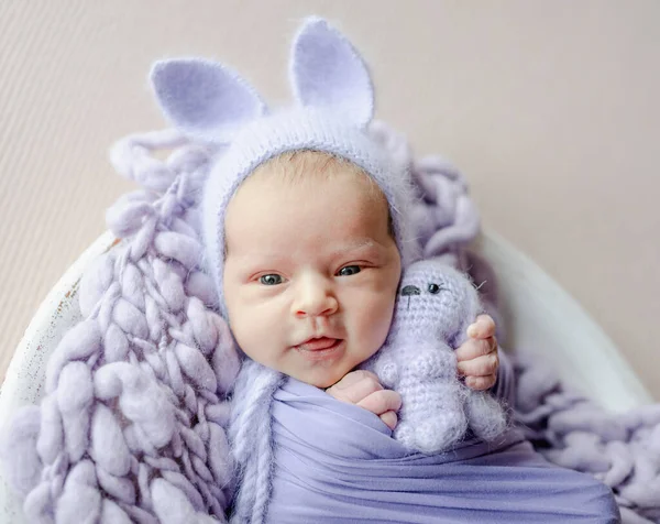 新生児の女の子ウサギの耳にウサギのおもちゃを保持し カメラを見てニット帽を着て 幼児子供嘘Swaddledでパープル生地スタジオポートレート — ストック写真
