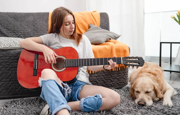 十几岁的女孩坐在地板上练习与金毛猎犬一起弹奏吉他 漂亮的吉他手 带着乐器 带着纯正的宠物狗 看着相机 — 图库照片