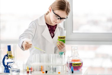Akıllı kız koruyucu gözlük takarak bilimsel kimya deneyi yapıyor ve şişeye ünvan tehlikesiyle birlikte sıvı ekliyor. Okul dersinde kimyasal teçhizatlı kız öğrenci.