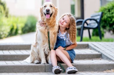 Küçük kız şapka takıyor Golden Retriever köpeğini kucaklıyor yazın dışarıda oturuyor. Güzel çocuk şehirde pofuduk köpekleri okşuyor.