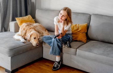 Golden Retriever köpeği olan genç bir kız elinde uzaktan kumandayla evdeki kanepede oturuyor. Labrador köpekli güzel çocuk televizyon kanalında çizgi film izliyor.