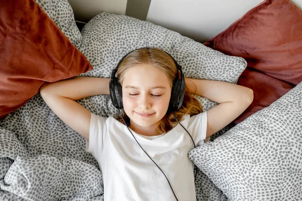 ヘッドフォンで美しい女の子の子供がベッドに横たわって 自宅で音楽を聞いています 美しい女性の子供休憩とともにイヤホンでベッドルームと笑顔 — ストック写真