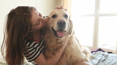 Güzel kız Golden Retriever köpeğine sarılıyor ve yatakta oturmuş gülümsüyor. Saf köpek labrador 'u olan genç bir kız evde kameraya bakıyor.