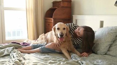 Güzel kız Golden Retriever köpeğine sarılıyor ve yatakta oturmuş gülümsüyor. Saf köpek labrador 'u olan genç bir kız evde kameraya bakıyor.