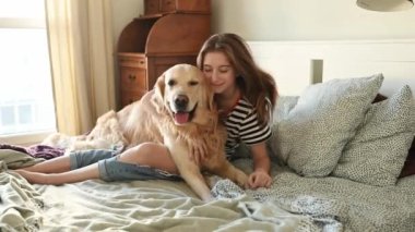 Güzel kız Golden Retriever köpeğine sarılıyor ve yatakta oturmuş gülümsüyor. Saf köpek labrador 'u olan genç bir kız evde kameraya bakıyor.