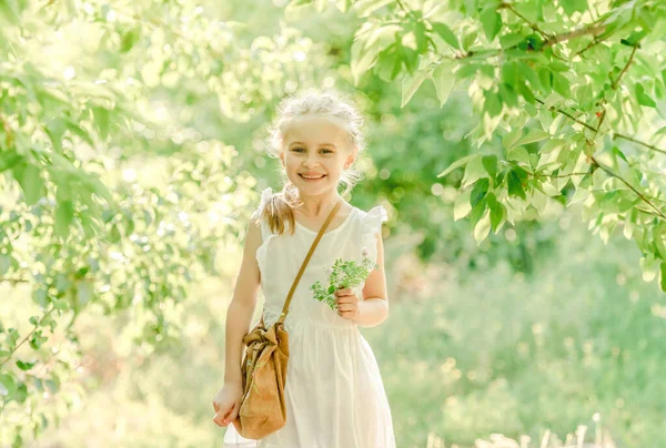 かわいい小さな女の子は 緑の公園で バッグと花を保持し 木々の間を歩いています — ストック写真