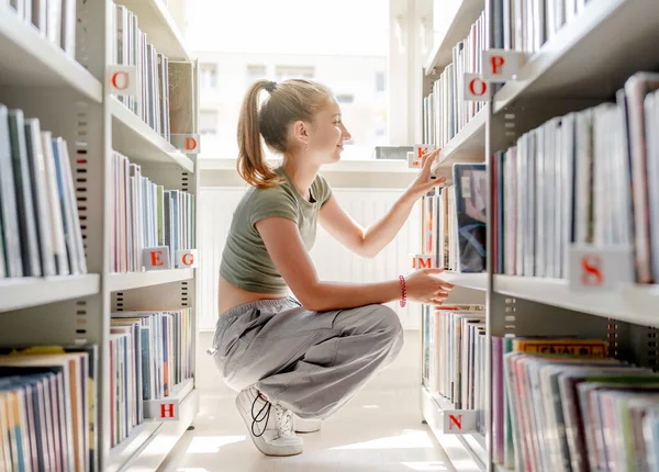 図書館の10代の少女が本棚で本を探している 美しい女性学生でカジュアル服を探している文学 — ストック写真