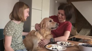 Altın av köpeği olan anne ve kızı kahvaltıyı hazırlıyor ve gülümsüyor. Safkan köpekli genç bir kadın ve kız ailesi kremalı süt yiyor.