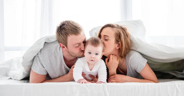 母と父は息子と毛布の下に横たわって 頬に赤ちゃんとキスをしました 美しい家族の朝一緒に日当たりの良いベッドルームで — ストック写真