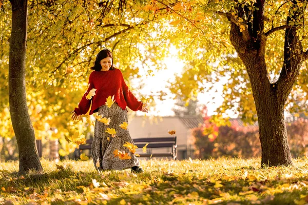 秋の公園に黄色の葉でポーズをとっているきれいな女の子 自然の中で黄金の葉と秋の季節に居心地の良い美しいドレスを身に着けている魅力的な若い女性 — ストック写真
