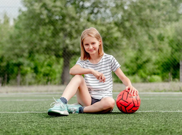 Pretty child girl sitting with football ball on grass and looking at camera. Cute female kid at socker field posing