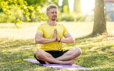 Yazın parkta paspasın üzerinde oturan bir adam yoga yapıyor. Adam kaslarını germek için pilates yapıyor.