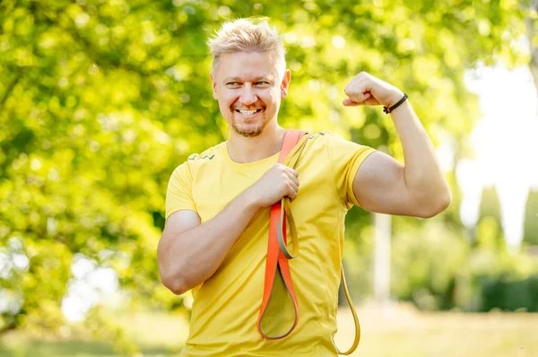 腕のトレーニングの後で筋肉を示す伸縮性があるゴム製バンドを持つ男 カメラと笑顔を見ているスポーツ用品を持つ男 — ストック写真